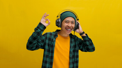 Smiling Asian man in a beanie and casual clothes gives an okay sign while listening to music or a podcast on his headphones. Isolated on a yellow background.