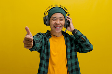 Asian man in a beanie and casual clothes smiles in approval while listening to music on his headphones. He gives a thumbs up gesture, seemingly enjoying the music. Isolated on a yellow background.