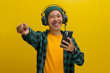 Asian man in a beanie and casual clothes points his finger directly at the camera while listening to music on his headphones. His phone is in his hand. Isolated on a yellow background.