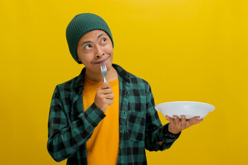 Pensive young Asian man wearing a beanie hat and casual shirt holds a spoon and an empty white...