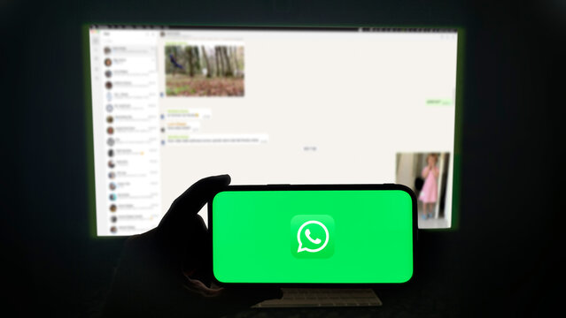 Whatsapp app running on a modern mobile phone. The smartphone is being held in landscape mode, in the darkness. A PC monitor with the Whatsapp desktop application in the background.