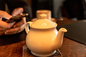 White teapot on the table, person holding smartphone in the dark background. Fiddle with a phone