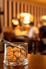 Pieces of brown sugar in a glass on wooden table, defocused people in a café or restaurant in the background. Organic demerara sugar