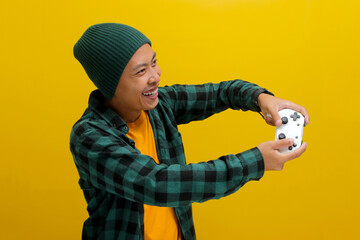 Side view of An enthusiastic Asian gamer, adorned in a beanie hat, casual shirt, and headphones, playing video games with a gamepad or joystick on a console, isolated on yellow background © Jamaludinyusup