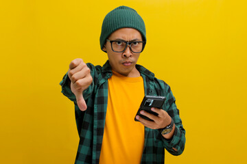 A smart young Asian man, wearing eyeglasses and dressed in a beanie hat and casual shirt gives a thumbs-down gesture while holding a phone, expressing disapproval, disagreement and giving a bad review