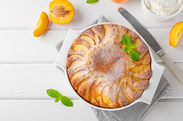 Delicious peach pie in a white ceramic baking form on a white wooden background. Summer cake. Selective focus, copy space.