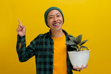 Asian man in a beanie and casual clothes smiles and points to the side at empty space, holding a...