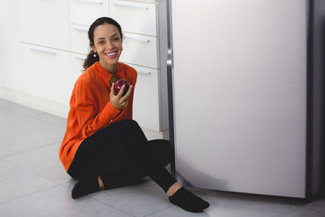 Beautiful African Latin American female is enjoy eating and holding a red apple from refrigerator,...
