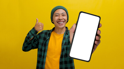 Excited young Asian man, dressed in a beanie hat and casual shirt, displays a blank white screen on...