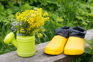 a watering can with flowers stands on a wooden bench and yellow galoshes next to it. Garden tools