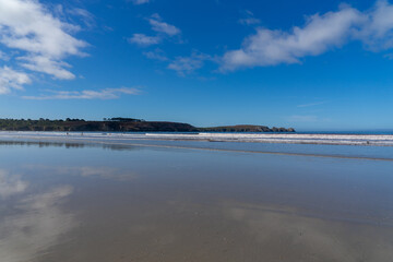 Reflets du ciel et nuages se miroitent sur le sable mouillé d'une plage de la presqu'île de Crozon, avec les falaises en arrière-plan, offrant une scène à la fois calme et majestueuse.