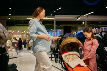 Pregnant mother and little daughter choosing stroller in shopping mall