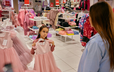 Charming little girl choosing elegant dress at shopping mall