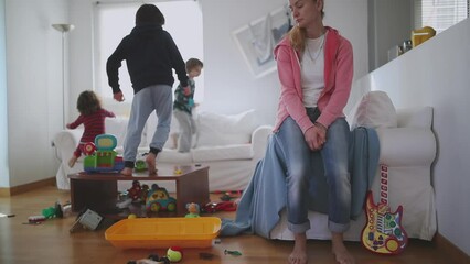 Exhausted mother sits with eyes closed and lying back on a couch, surrounded by her playful children and a disheveled living room strewn with toys, slow motion.