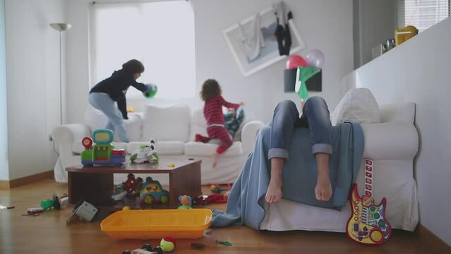 Exhausted mother sits with eyes closed and lying back on a couch, surrounded by her playful children and a disheveled living room strewn with toys, slow motion.