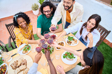 Top view of cheerful group young friends toasting glasses red wine celebrating rooftop BBQ party. Millennial people together enjoying alcoholic beverages sitting at terrace lunch table outdoors