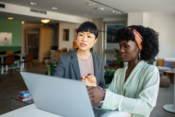 Two young businesswomen discussing work over laptop in office