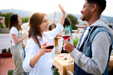 Excited multiracial young couple celebrating rooftop party drinking red wine. Friends dancing at...