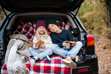 Cute child girl and boy 4-5 year old wearing knitted sweater and hat eating biscuits in car trunk outdoor in forest. Autumn season. Childhood.