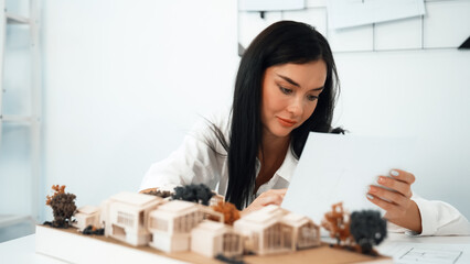 Closeup of young beautiful architect compares blueprints with architectural model with house model...