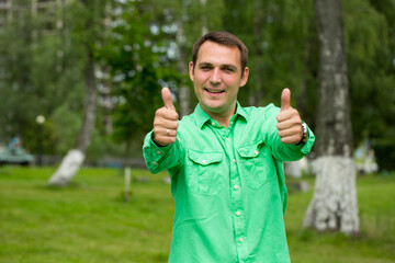 Young handsome man with thumb up in a green shirt on the background of summer park