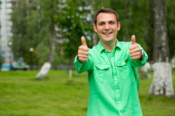 Young handsome man with thumb up in a green shirt on the background of summer park