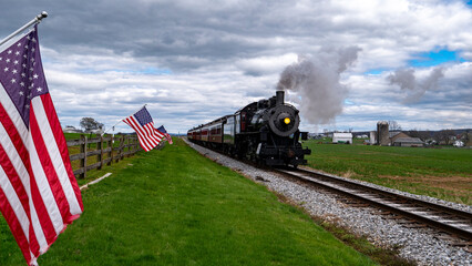 A train is traveling down the tracks next to a field with American flags. The train is black and white and is pulling a passenger car. The flags are waving in the wind