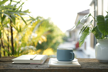 Blue coffee cup on wooden tray and notebook and Philodendron plant on wooden table under sunlight
