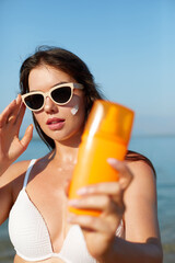 Beautiful young woman with sun cream on face. Girl holding sunscreen bottle on the beach and...