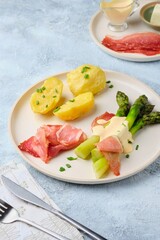 Green boiled asparagus served with boiled potatoes, thinly sliced smoked pork and sauce on a round clay plate on a gray concrete background. Asparagus recipes.
