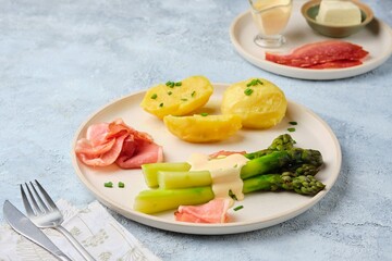 Green boiled asparagus served with boiled potatoes, thinly sliced smoked pork and sauce on a round clay plate on a gray concrete background. Asparagus recipes.