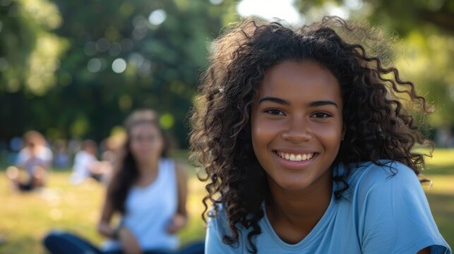 A young woman with curly hair clad in a blue t shirt flashes a radiant smile as she poses for the camera in the park with her friends leisurely seated in the background blurred out enjoying