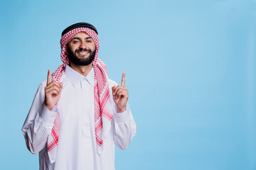 Excited muslim sales person pointing upwards direction while posing for studio portrait. Cheerful smiling arab man wearing traditional clothes showing up and looking at camera