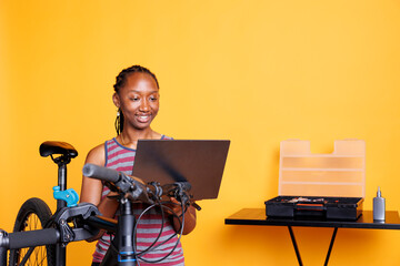 African American female cyclist repairs broken bike utilizing toolbox and personal computer to...