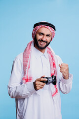 Excited muslim man holding joystick and showing clenched fist while winning game studio portrait....