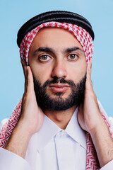 Man wearing muslim checkered headscarf covering ears with arms and looking at camera closeup. Arab...