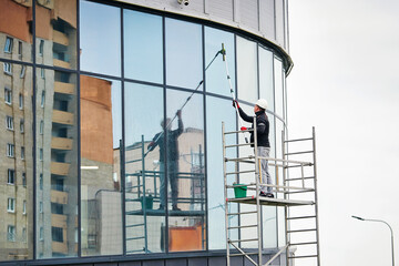 Worker on scaffold washes glass facade of business center. Window washer wipe wet windows at...