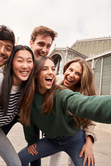 Fototapeta premium Vertical. Group happy young people smiling making gestures while taking selfie together at travel event. Generation z friends having fun and sharing positive moment posing cheerful for photo outdoors