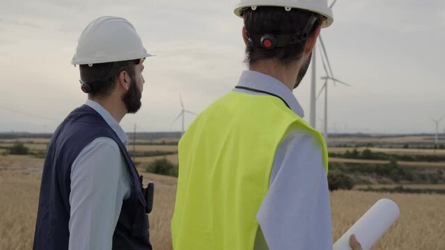 Engineers worker group wind power plant walking together planning of mill wearing yellow vest and helmet, equipped working tools at windmill farm. Professional technicians men