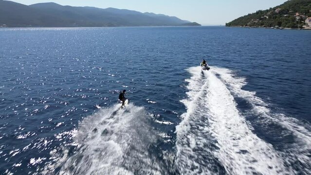Aerial drone footage of a person water skiing behind a speedboat in a vast blue sea, with scenic coastline and hills in the background.