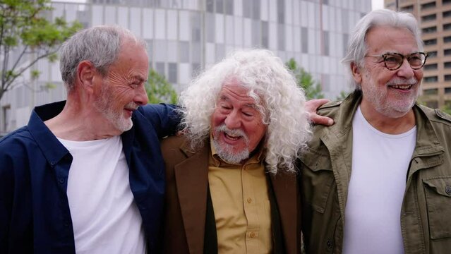 Group of three mature caucasian men enjoying embracing walk together laughing outdoor. Senior male old friends having fun hugging strolling happy in the urban city street. Elderly people golden ager