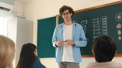 Professional mentor looking around classroom while standing in front of blackboard with math formula or theory. Diverse elementary student learning or studying and sitting in Math lesson. Pedagogy.