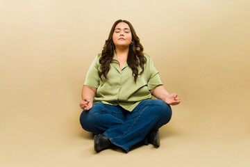 Calm plus-size woman meditates in lotus pose with a serene expression in a studio setting