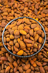 Saffron Almonds in a Bowl Surrounded by Loose Almonds, A close-up view captures saffron almonds piled in a metal bowl, surrounded by a sea of loose almonds. The image is vibrant and detailed.