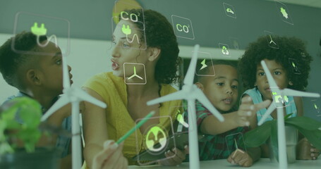 Image of energy icons over happy diverse female teacher and pupils with wind turbines in class - Powered by Adobe