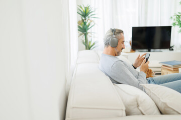 Man in headphones focused on smartphone with chihuahua dog on couch, spacious living room with TV...