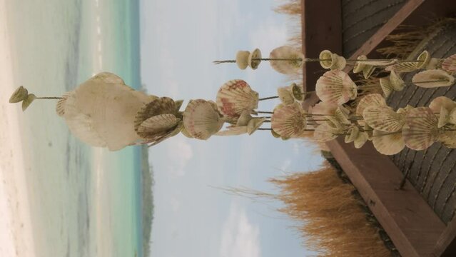 Pendant seashells swinging on background of sandy ocean beach hanging from roof of thatched canopy on tropical island, vertical video.