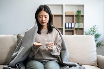 Young Asian woman suffering from flu symptoms, covered with a blanket on a sofa. Concept of...