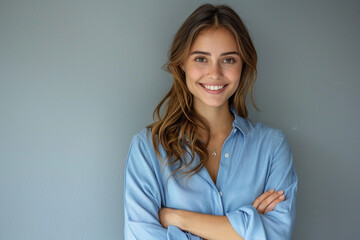 Obraz premium A confident, smiling businesswoman in a blue shirt, arms crossed, exudes professionalism. Her stylish demeanor and positive energy shine on the gray backdrop.
