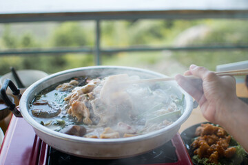 Slice of pork was picking up by the chopstick in the woman's hand above the curved brass pan around...